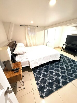 A hotel room with a bed, a TV, a desk with a remote, and a patterned rug.