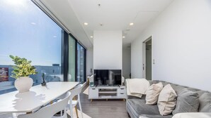 Een moderne woonkamer met een flatscreen televisie, een grijze bank en een eethoek met een witte tafel en stoelen.