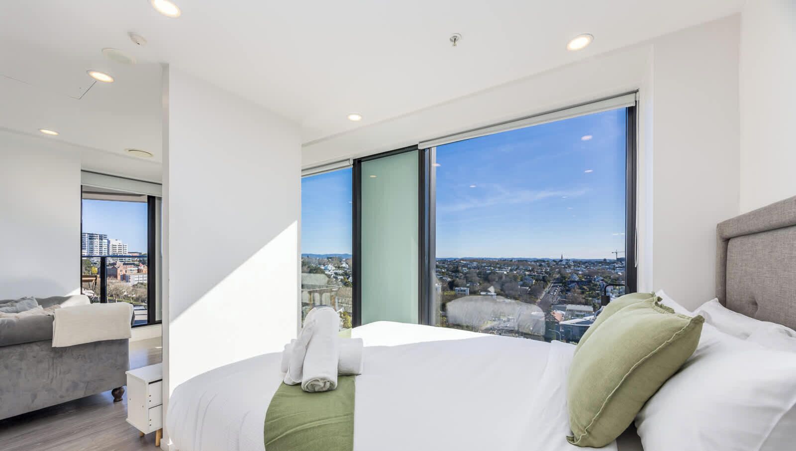 A modern hotel room with a large bed, a view of a cityscape through floor-to-ceiling windows, a grey sofa, and a small bedside table.