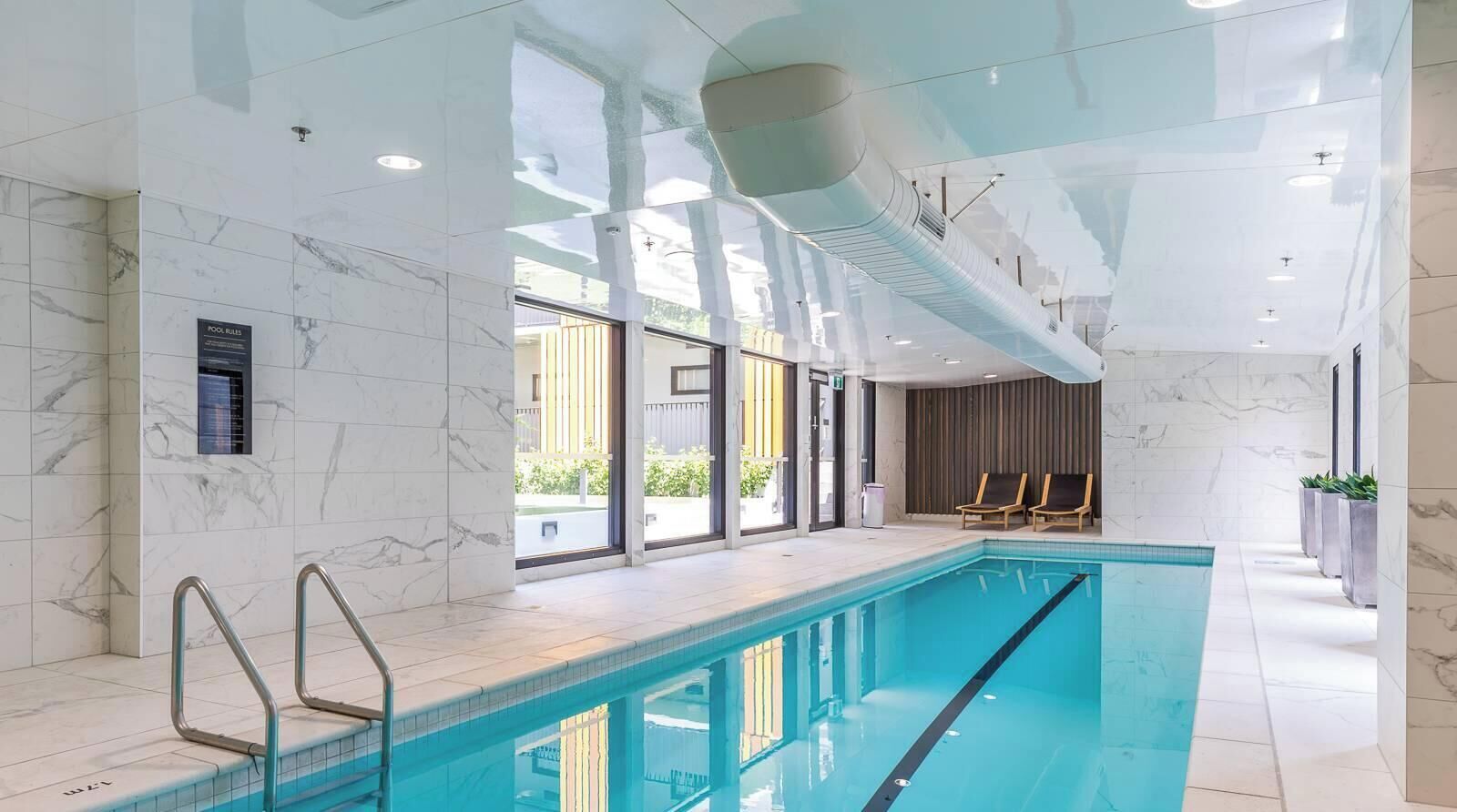 An indoor swimming pool with clear glass walls, a marble-tiled floor, and a ceiling with a central air vent.
