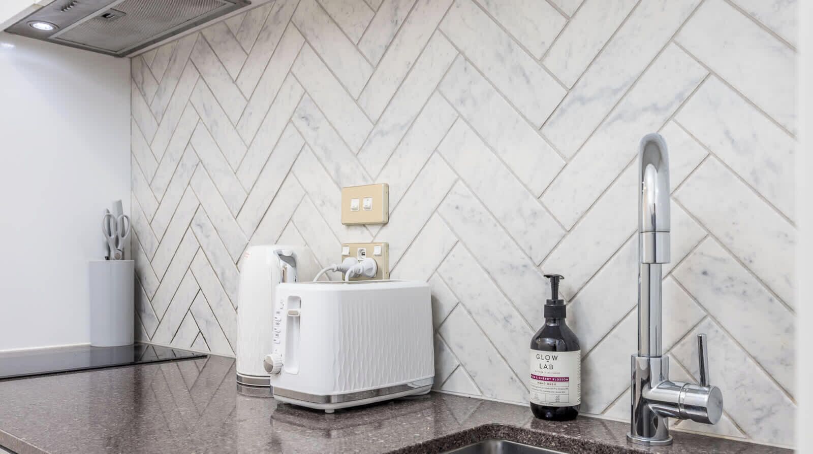 A kitchen with a herringbone tile backsplash, a toaster, a sink, and a bottle of Glow Lab hand soap.