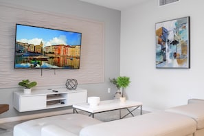 A modern living room with a flat-screen TV mounted on a textured wall, a white entertainment unit, a coffee table, and a sofa. - 2BD Prime Location & Free Resort Access! (Kissimmee)