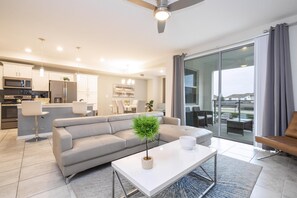 A modern living room with a sofa, coffee table, and a view of the city through large windows. - 2BD Prime Location & Free Resort Access! (Kissimmee)