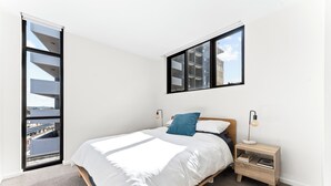 Ein Schlafzimmer mit einem Bett, einem Nachttisch, einer Lampe und einem Fenster mit Blick auf Gebäude.