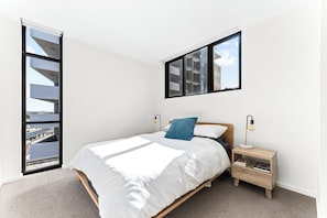 A bedroom with a bed, a nightstand, a lamp, and a window with a view of buildings.