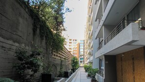 Um beco estreito entre edifícios, com uma parede de concreto de um lado e um edifício moderno do outro. Há plantas em vasos ao longo do beco e uma escadaria que leva ao prédio.