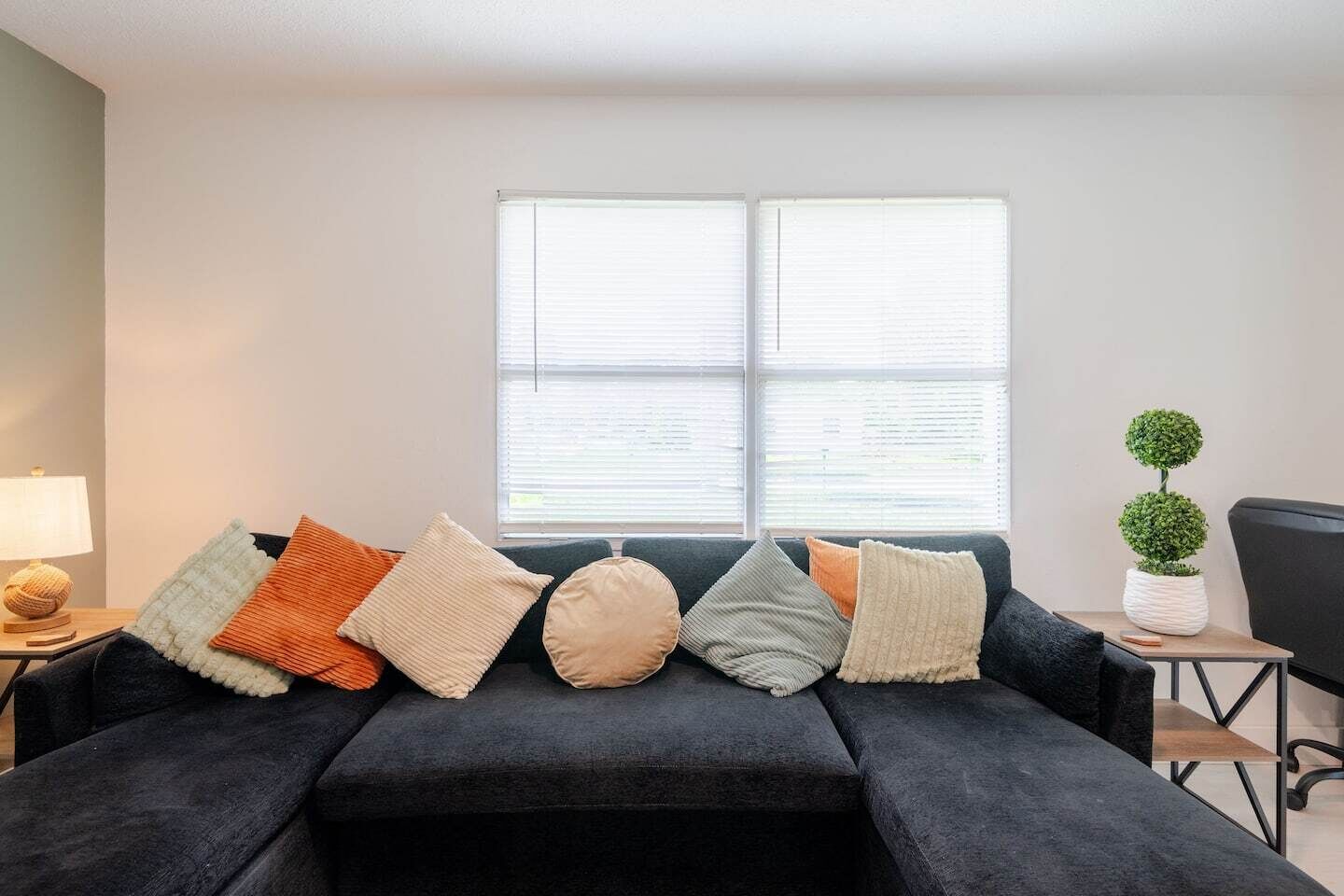 A modern living room with a dark sofa, multiple cushions, a lamp, a small table with a plant, and a window with blinds.