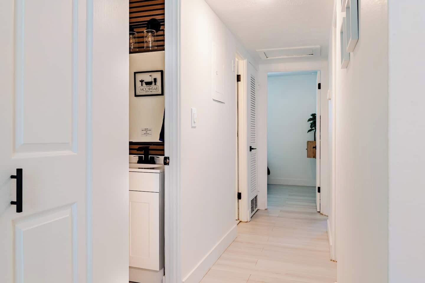 Un couloir avec une porte blanche, une salle de bain avec un lavabo et un miroir, et une plante au loin.