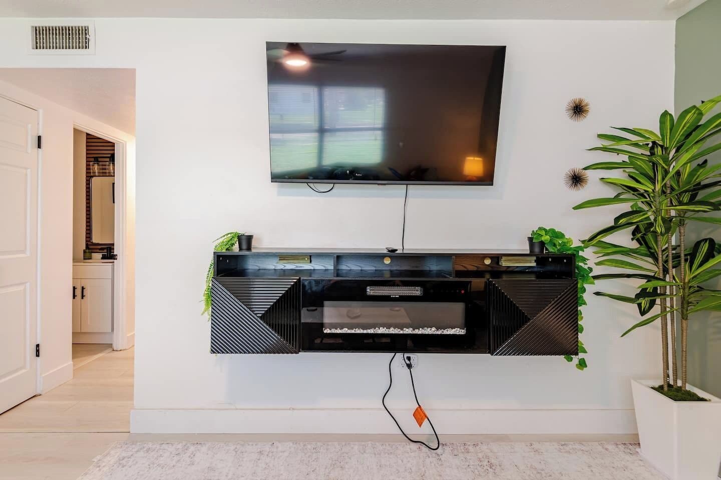 A modern living room with a flat-screen TV mounted on the wall, a black entertainment unit below, and a potted plant to the right.