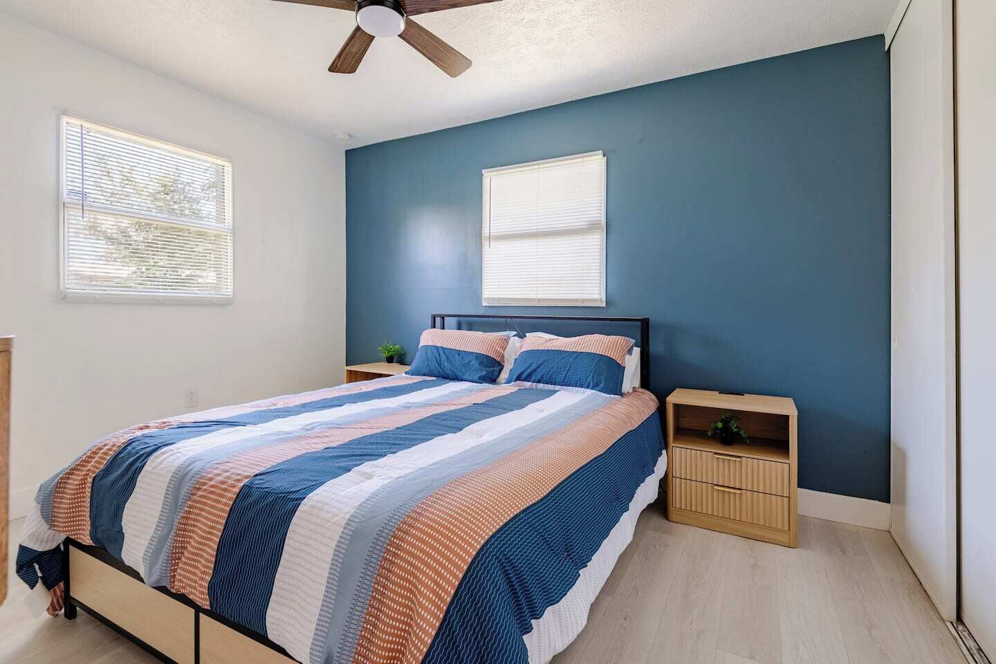 A bedroom with a bed, two bedside tables, a ceiling fan, and two windows with blinds.