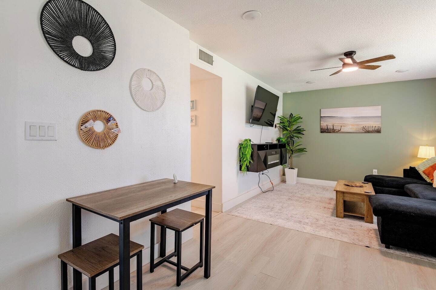A modern living room with a dining area, a flat-screen TV mounted on the wall, a ceiling fan, and a painting on the wall.