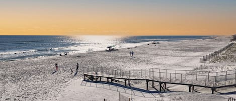 Una playa con un paseo marítimo de madera que lleva al agua, terreno arenoso y cielo despejado.