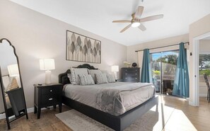 Ein Schlafzimmer mit einem Bett, Nachttischen, einer Kommode, einem Deckenventilator und einem großen Fenster mit Vorhängen.