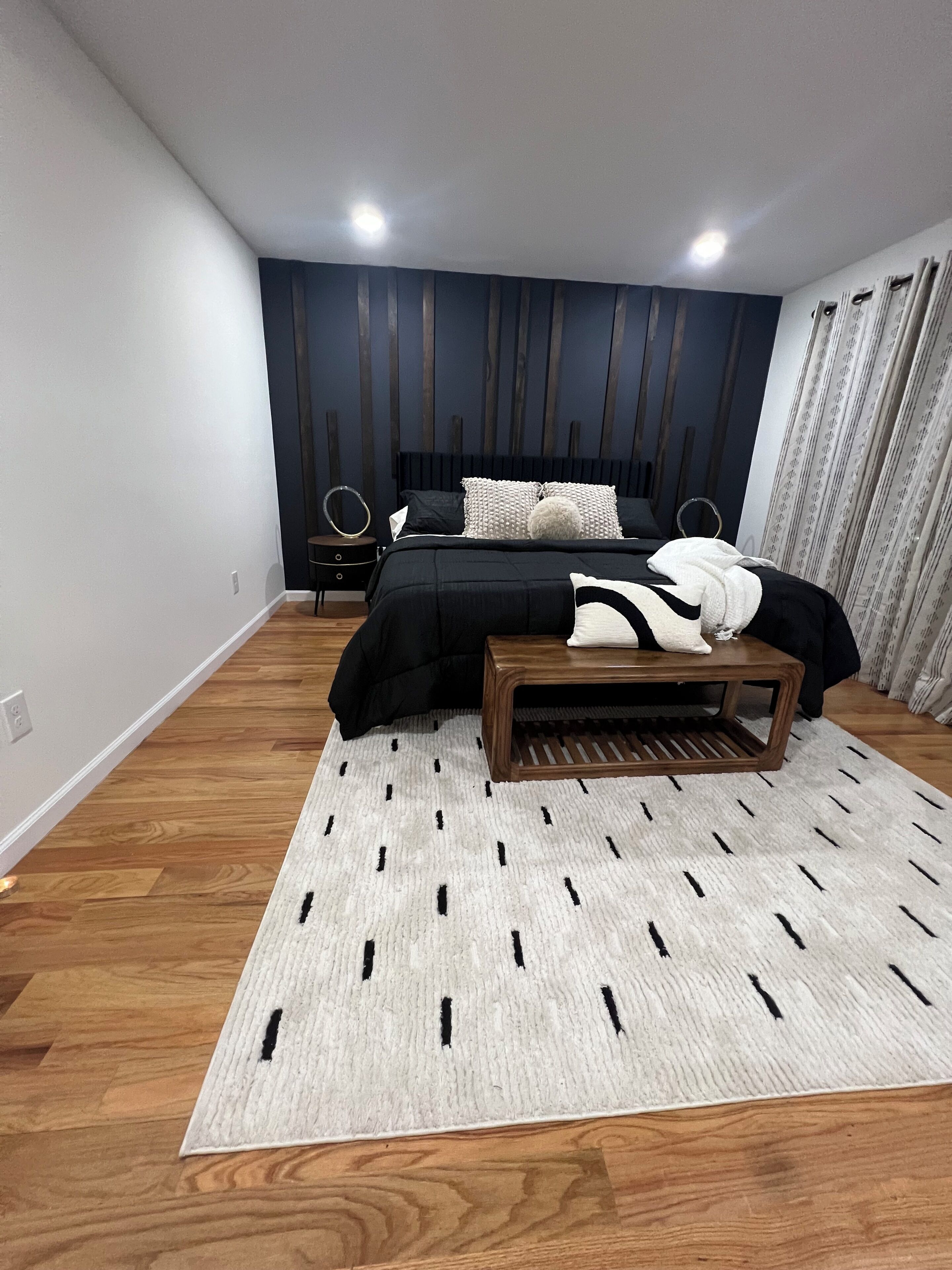 Een moderne slaapkamer met een groot bed, een nachtkastje, een houten bankje en een kamerkleed met een patroon.
