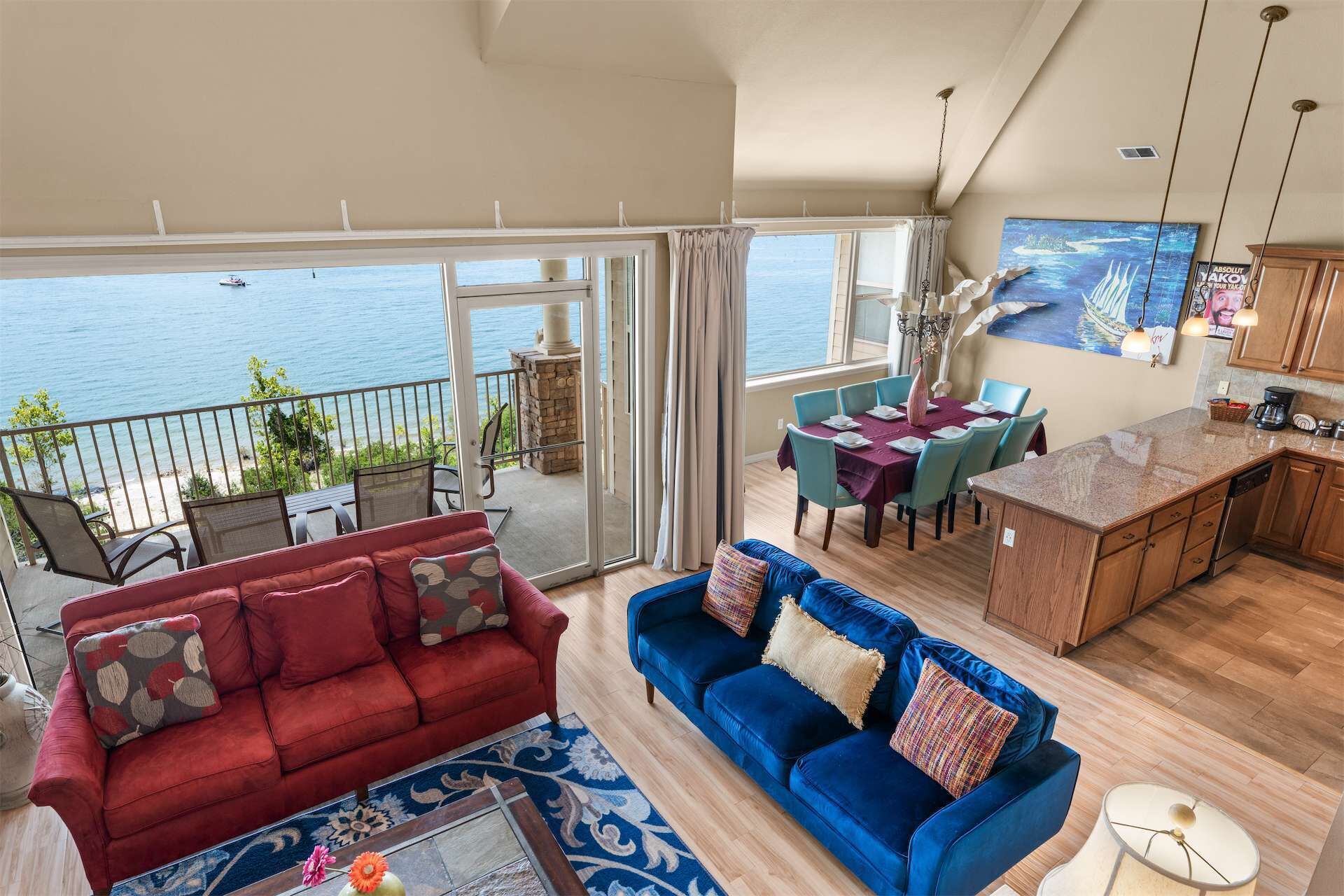 Ein geräumiges Wohnzimmer mit einer roten und blauen Sofagarnitur, einem Esstisch mit Stühlen, einer Küchenzeile und Meerblick durch große Fenster.