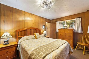 Ein Schlafzimmer mit Holzkopfende, einem Bett mit gemusterten Textilien, einem Schminktisch, einem Stuhl und einem Fenster mit Vorhängen.