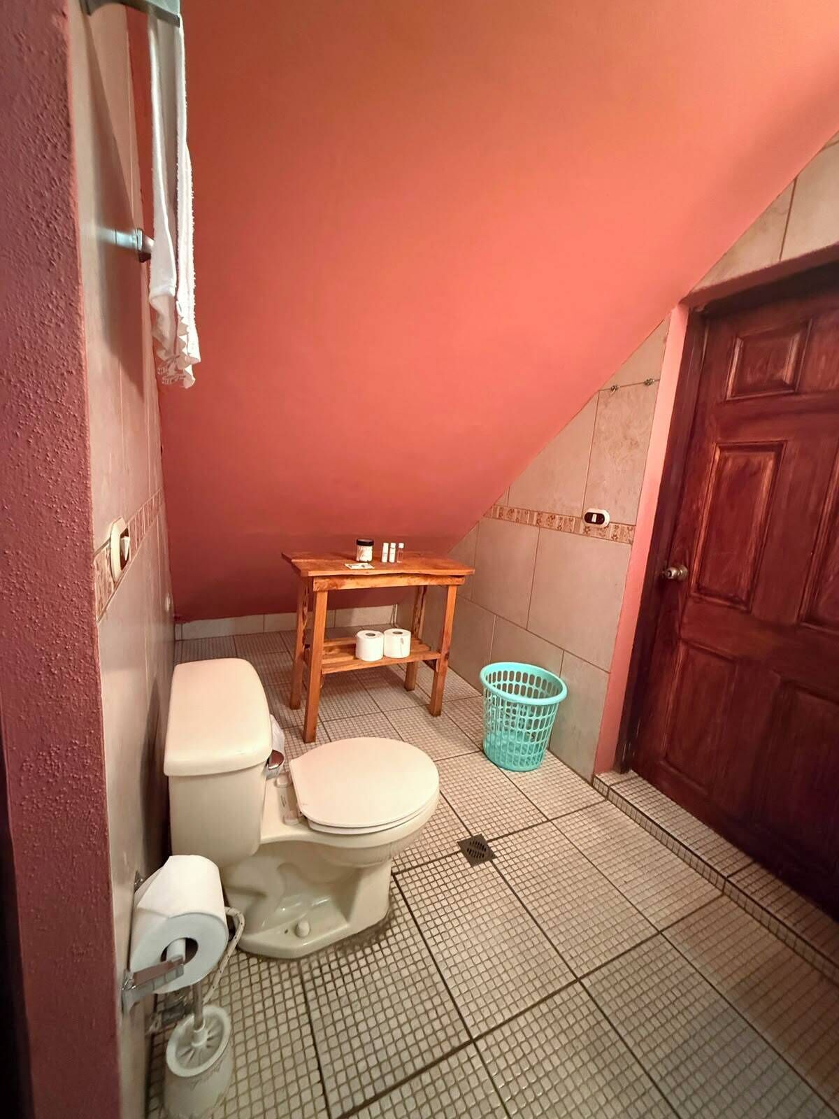 A small bathroom with a toilet, a wooden shelf with toiletries, a blue laundry basket, and a wooden door.