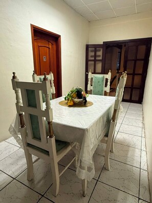 A dining area with a table set for four, surrounded by chairs with wooden frames and cushioned seats. A basket with fruits and a decorative item is placed on the table.