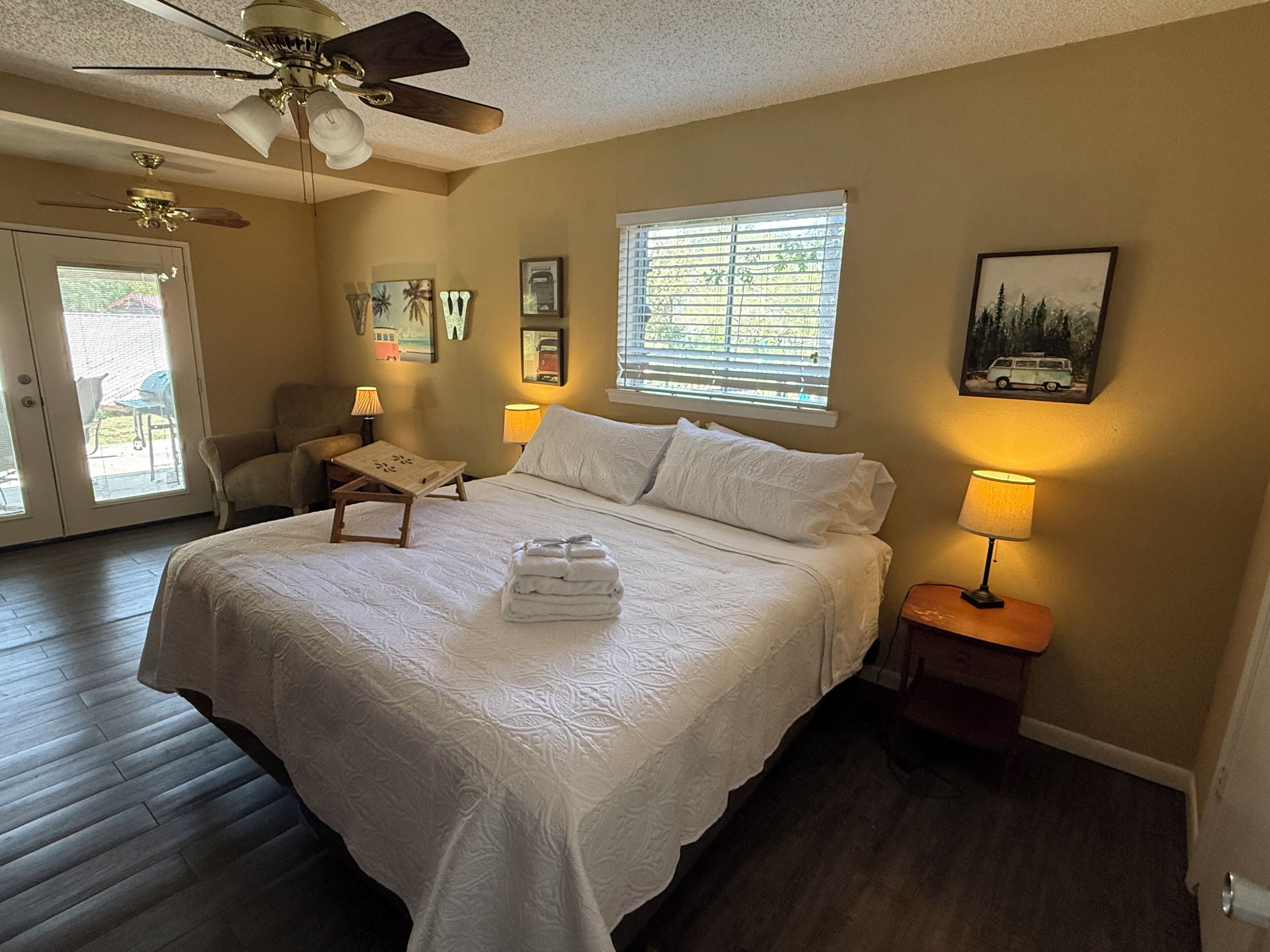 A bedroom with a bed, a ceiling fan, a window with blinds, a chair, a table, and a lamp.