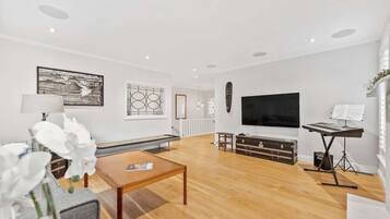 A spacious living room with a wooden floor, a flat-screen TV, a music stand, and a piano.
