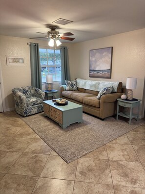 A living room with a beige couch, patterned armchair, and a coffee table. There is a ceiling fan, a window with curtains, and a painting on the wall. - Quaint 3 bedroom 2 bath house centrally located in Saint Petersburg (Pinellas Park)