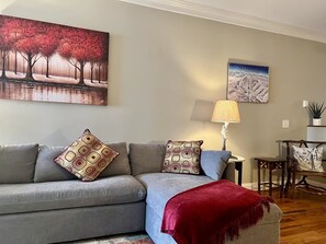 Ein Wohnzimmer mit grauem Sofa, dekorativen Kissen mit Muster, einer roten Decke, einer Lampe und zwei an der Wand hängenden Bildern.