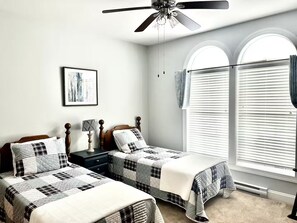 Ein Schlafzimmer mit zwei Betten, einem Deckenventilator, einem Nachttisch, einer Lampe, einem gerahmten Bild und einem Fenster mit Jalousien.