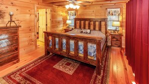 A wooden cabin bedroom with a bed, a nightstand, a dresser, a ceiling fan, and a window with curtains. - Game Room  / Mtn Views  / Hot Tub / Fire Pit & Fire Table /  Coffee Bar / (Lake Lure)