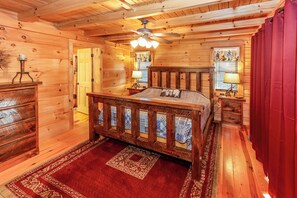 A wooden cabin bedroom with a bed, a nightstand, a dresser, a ceiling fan, and a window with curtains. - Game Room  / Mtn Views  / Hot Tub / Fire Pit & Fire Table /  Coffee Bar / (Lake Lure)