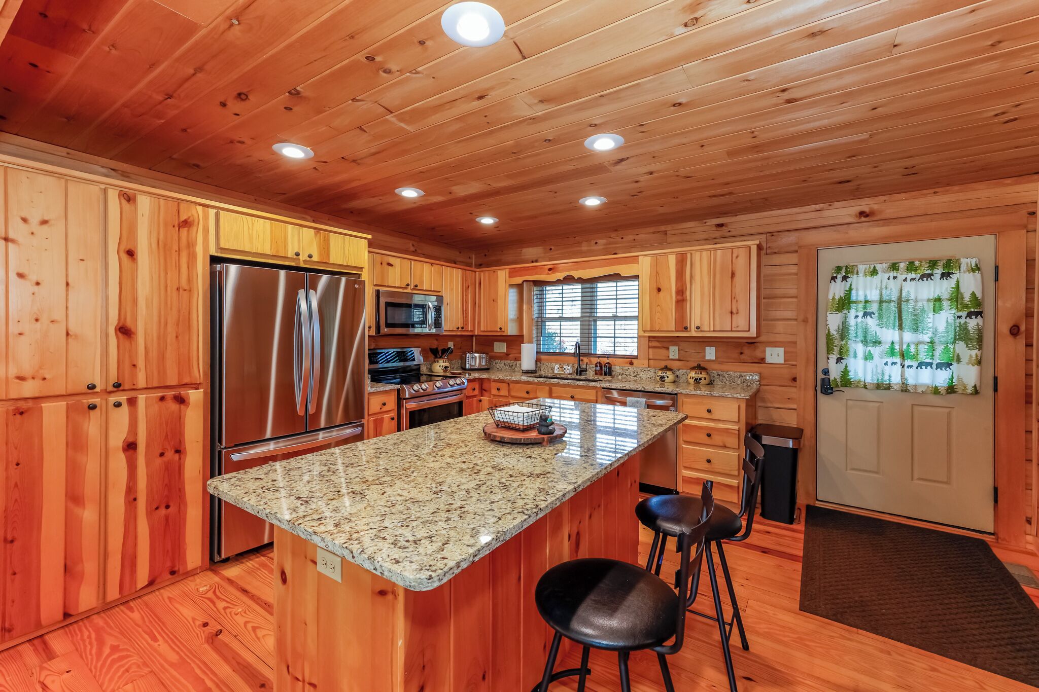 A wooden cabin interior with a kitchen featuring a stainless steel refrigerator, a microwave, and a granite countertop.