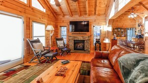 A log cabin interior with a fireplace, leather sofa, and a flat-screen TV. - Game Room  / Mtn Views  / Hot Tub / Fire Pit & Fire Table /  Coffee Bar / (Lake Lure)