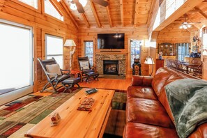 A log cabin interior with a fireplace, leather sofa, and a flat-screen TV. - Game Room  / Mtn Views  / Hot Tub / Fire Pit & Fire Table /  Coffee Bar / (Lake Lure)