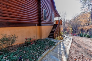 Une cabane en rondins avec une façade en bois, un chemin de pierre menant à un escalier en bois, et un jardin agrémenté de diverses plantes et d’une chaise rouge.