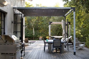 Une terrasse extérieure équipée d’un barbecue, d’une table à manger, de chaises et d’une pergola offrant de l’ombre.
