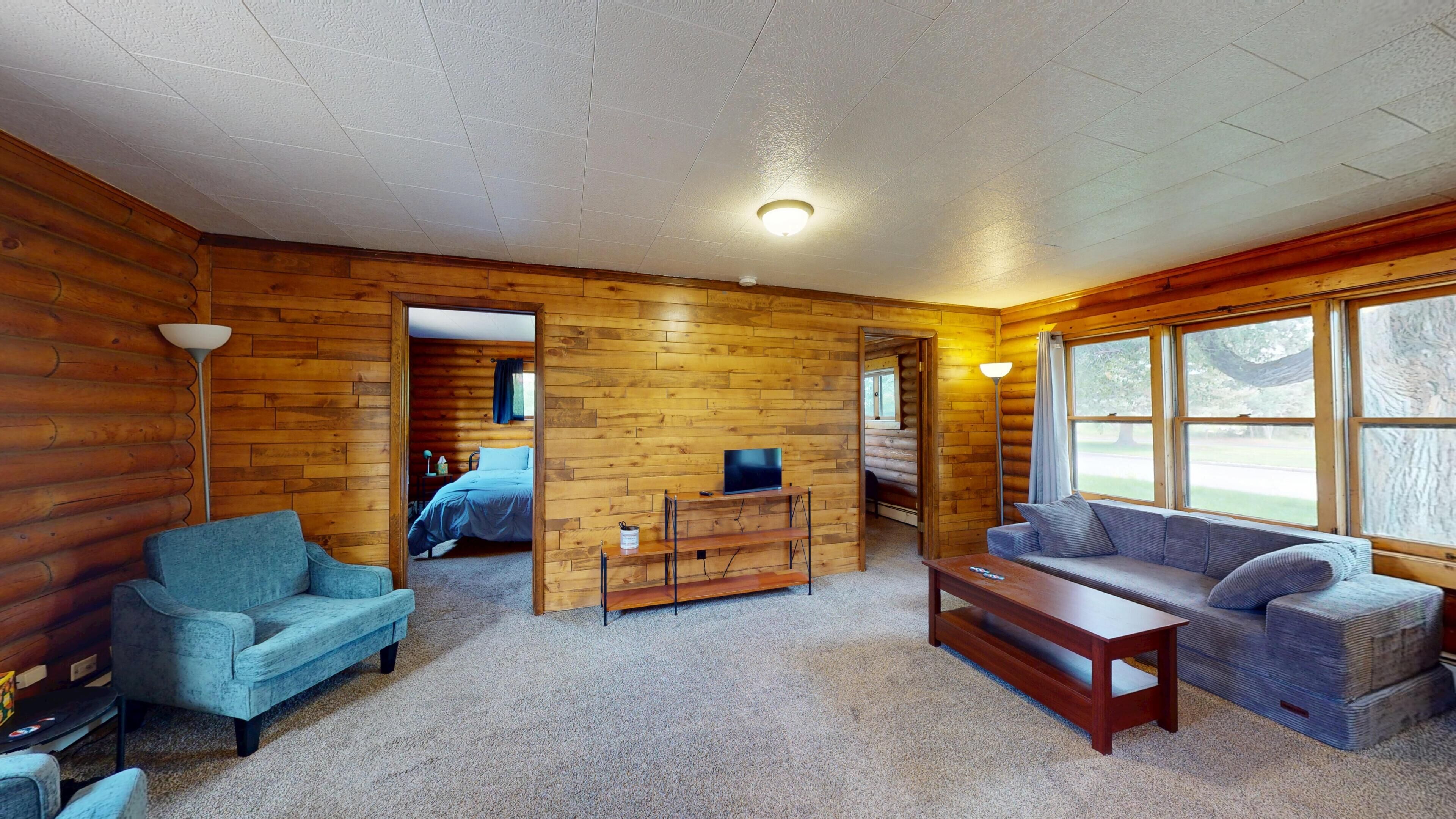 A log cabin interior with a sofa, a television on a stand, a lamp, and a window with a view.