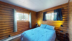 A log cabin bedroom with a bed, two bedside lamps, and a window with curtains. - Gunni Getaway - Pet Friendly (Gunnison)