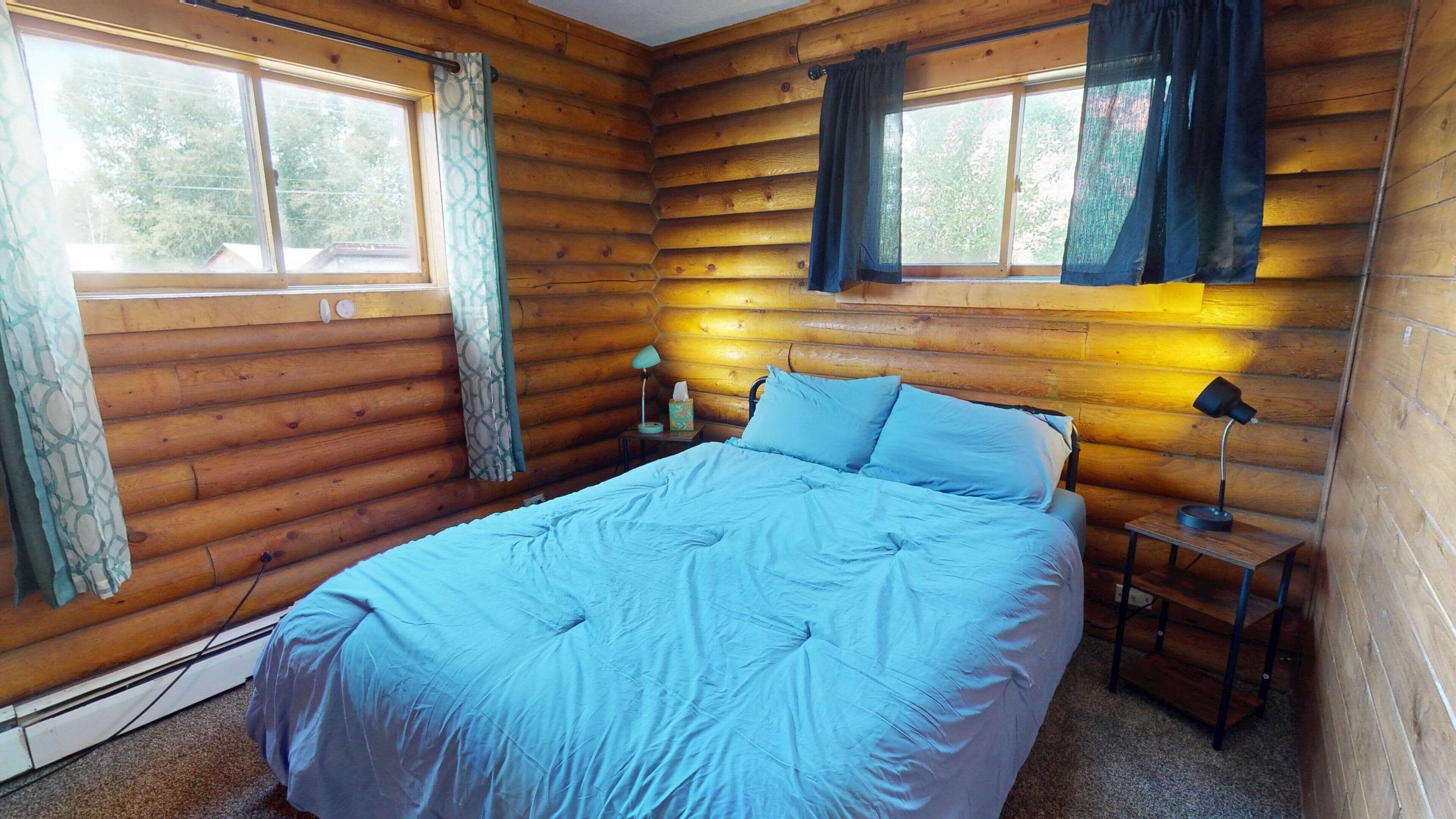A log cabin bedroom with a bed, two bedside lamps, a nightstand, and curtains.