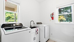 A laundry area with a Samsung washer and dryer, a window with a view of trees, and a fire extinguisher mounted on the wall. - Monthly Stay | Vineyard Pool, Hot Tub | 3BR Home | Sleeps 6, Pets OK, West Dry Creek (Healdsburg)