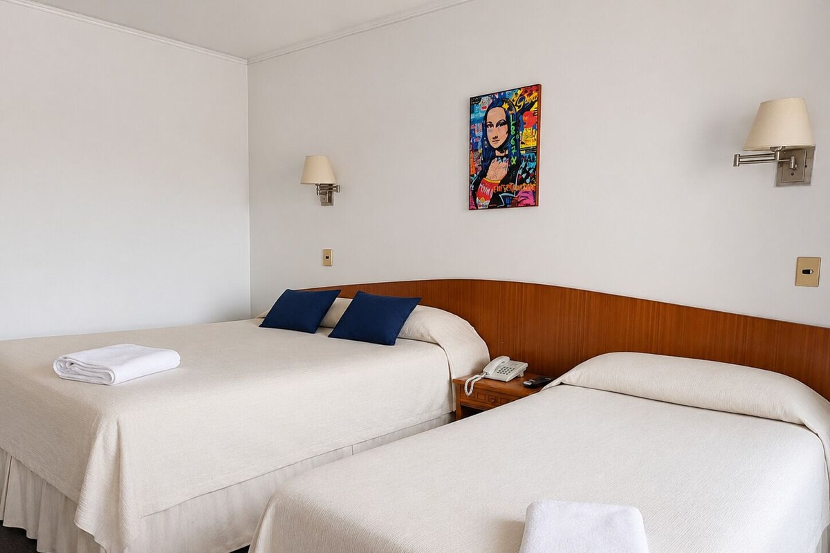 Habitación de hotel con dos camas, cabecero de madera, mesita de noche con lámpara, teléfono y una obra de arte colorida en la pared.