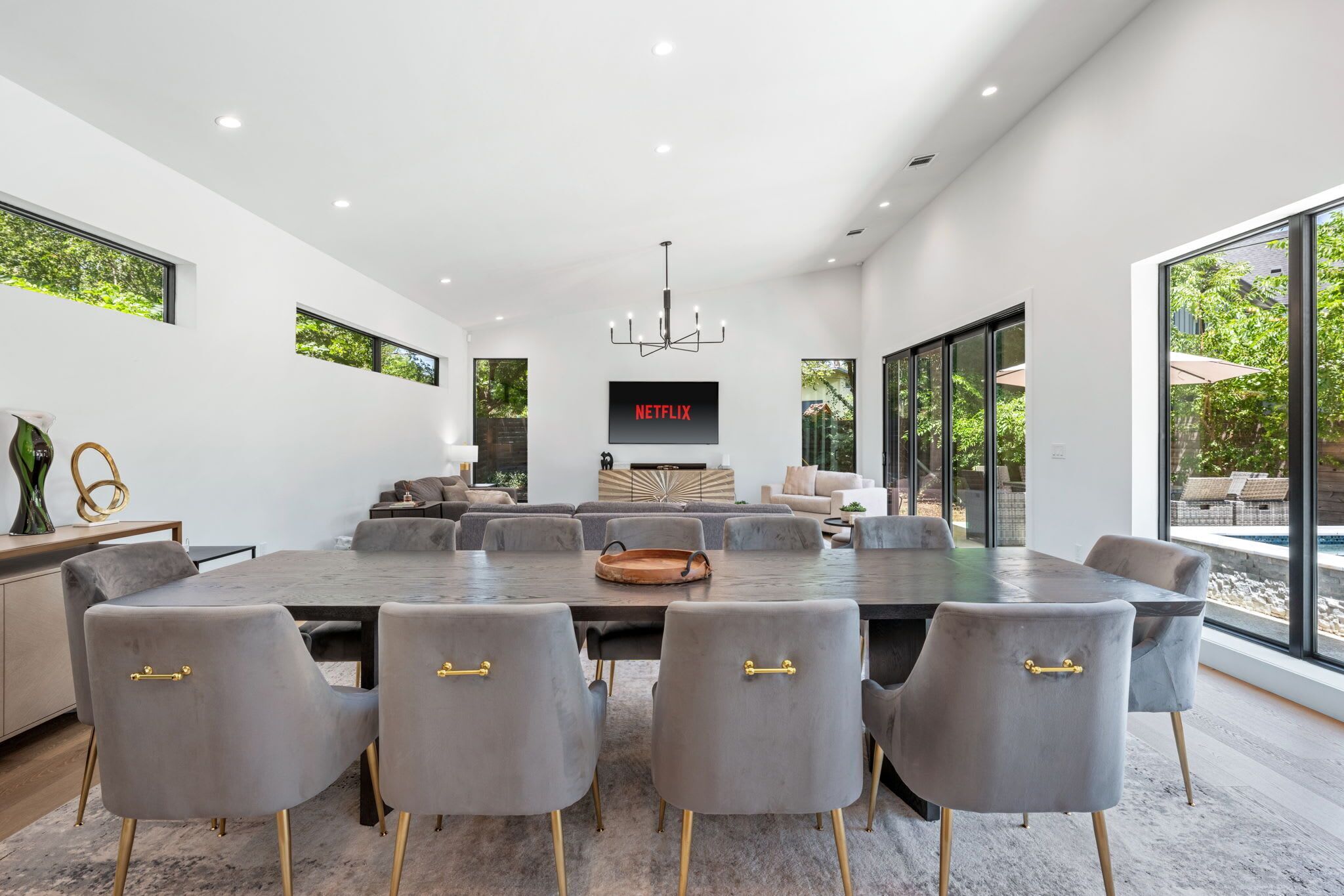 A modern dining room with a large table, grey upholstered chairs with gold handles, a chandelier, and a television screen displaying 'NETFLIX'.