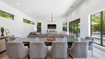 A modern dining room with a large table, grey upholstered chairs with gold handles, a chandelier, and a television screen displaying 'NETFLIX'.