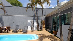 A swimming pool with lounge chairs and a table, surrounded by palm trees and a modern building with large windows. - Edícula 55 M2 aceita pequenos pets (Foz do Iguaçu)