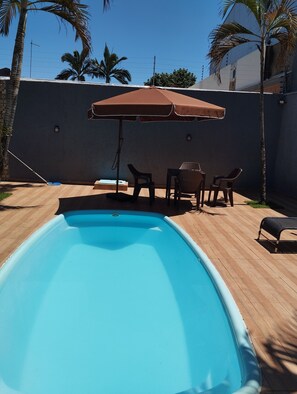 A swimming pool with a wooden deck, a table with chairs, and a large umbrella.