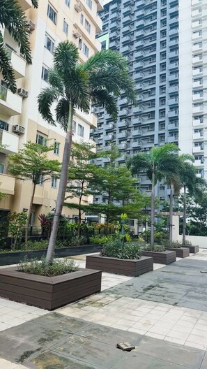 A residential area with modern apartment buildings, a paved walkway, and landscaped planters with various shrubs and trees. - Condo in Araneta City Cubao • Walk to Coliseum • Smart Lock • Parking • Pool (Quezon City)