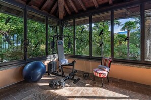 A gym with exercise equipment, a blue exercise ball, and a chair, with large windows overlooking greenery. - Villa Donzella - Luxury historic villa with pool (Menaggio)
