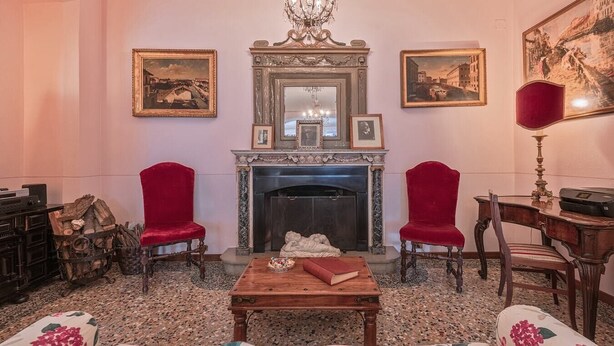 Een kamer met een open haard, twee rode fauteuils, een houten tafel met een boek, en lijstjes met afbeeldingen aan de muren.