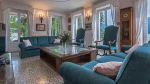 Een ruime kamer met een blauwgroene zithoek, een houten salontafel en een groot raam met gordijnen.