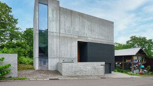 Un edificio moderno con un amplio ventanal de vidrio, fachada de concreto y una estructura de madera contigua.