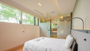 Habitación de hotel moderna con una cama grande, cabina de ducha de vidrio y vistas a árboles por la ventana.