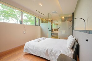 Ein modernes Hotelzimmer mit einem großen Bett, einer Duschkabine aus Glas und Blick auf Bäume durch das Fenster.